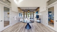 Located at Windsor Cay Blvd in Clermont, Florida, this welcoming entrance lobby showcases a thoughtfully designed single-floor layout that blends modern coastal aesthetics with functional communal space. The entrance greets visitors through glass double doors framed by floor-to-ceiling patterned drapery and transom windows, flooding the foyer with natural light and offering views of the surrounding community.

The soaring ceilings in the entry zone create an airy, grand first impression, accentuated by crisp white shiplap wall paneling and exposed dark wood beams that add warmth and architectural contrast. Light-toned wood-look flooring runs throughout, tying the space together seamlessly. A sculptural dark round pedestal table anchors the center of the entrance area, complemented by neutral-toned upholstered seating for guests.

Transitioning deeper into the space, a stylishly appointed lounge area features a diagonal shiplap accent wall, a wall-mounted flat-screen television, and a white built-in credenza with dual beverage refrigerators—ideal for entertaining or casual gatherings. A pendant light fixture and recessed lighting provide layered illumination throughout. Glass-paned interior French doors connect to adjacent rooms, enhancing the sense of openness. The overall ambiance is refined yet relaxed, reflecting a contemporary Florida lifestyle.