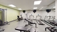 Located at 15 Eva Rd in Toronto, Ontario, this fitness room offers a well-organized exercise space designed to support a variety of workout routines. The room features a clean, functional layout with durable speckled flooring and neutral-toned walls accented by a fresh green feature wall, creating an energizing atmosphere.

The space is equipped with a comprehensive selection of cardio machines, including multiple treadmills, an elliptical trainer, and both upright and recumbent stationary bikes. For strength training, a dumbbell rack with a full range of free weights, a multi-purpose adjustable weight bench, and a cable machine are conveniently arranged throughout the room. Exercise balls in multiple sizes are stored on a dedicated rack near the entrance, adding versatility for core and flexibility training.

Overhead, recessed flat-panel lighting provides bright, even illumination throughout the space. A wall-mounted split air conditioning unit ensures a comfortable temperature year-round. Wall-mounted televisions are positioned for easy viewing during cardio sessions. A large mirror along one wall enhances the sense of openness and allows users to monitor their form during workouts. Posted fitness area rules and informational signage reflect a well-managed facility. The room is accessed through a secure door with a glazed panel, ensuring both privacy and safety for users.