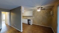 This single-floor residence offers a practical and well-organized layout designed for comfortable everyday living. The open-concept design seamlessly connects the living area to the kitchen, creating a sense of spaciousness and ease of movement throughout the main living space.

The kitchen is outfitted with white cabinetry that provides ample storage, a stainless steel dishwasher, and a single-basin sink set into a light-toned countertop. The cabinetry spans both upper and lower sections, maximizing functionality. The kitchen's warm, neutral wall color complements the crisp white of the cabinets, creating a clean and inviting cooking environment.

The living area features rich, dark hardwood-style flooring that runs throughout, adding warmth and visual continuity to the space. A ceiling fan with an integrated light fixture is centrally positioned, offering both ambient lighting and air circulation. The walls are finished in a calming gray-taupe tone, contributing to a neutral and versatile palette that adapts well to a variety of furnishing styles.

A textured popcorn ceiling adds a classic touch overhead, while white baseboards and trim provide clean, defined borders throughout the space. A partial wall divides the kitchen from the living area, offering a subtle sense of separation while maintaining the open feel of the overall layout.