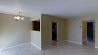 This single-floor residence offers a clean, functional layout with a cohesive design throughout. The main living area is spacious and open, featuring light-colored ceramic tile flooring and neutral beige walls that create a bright, welcoming atmosphere. A central half-wall partition with a pass-through window connects the living space to the kitchen, allowing for an open feel while maintaining a defined separation between the two areas. The kitchen is equipped with dark wood cabinetry, providing a rich contrast against the lighter tones of the surrounding walls and flooring. A doorway adjacent to the kitchen partition leads further into the home, offering access to additional rooms. Overhead, the ceilings are smooth and white, contributing to the sense of openness in the space. A pendant light fixture adds a subtle decorative touch near the kitchen pass-through. HVAC vents are integrated into the ceiling and walls, ensuring climate comfort throughout. White baseboards run along the perimeter of the rooms, adding a crisp, finished look. Overall, the space presents a straightforward and practical layout suited for comfortable everyday living.