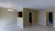 This single-floor residence offers a clean, functional layout with a cohesive design throughout. The main living area is spacious and open, featuring light-colored ceramic tile flooring and neutral beige walls that create a bright, welcoming atmosphere. A central half-wall partition with a pass-through window connects the living space to the kitchen, allowing for an open feel while maintaining a defined separation between the two areas. The kitchen is equipped with dark wood cabinetry, providing a rich contrast against the lighter tones of the surrounding walls and flooring. A doorway adjacent to the kitchen partition leads further into the home, offering access to additional rooms. Overhead, the ceilings are smooth and white, contributing to the sense of openness in the space. A pendant light fixture adds a subtle decorative touch near the kitchen pass-through. HVAC vents are integrated into the ceiling and walls, ensuring climate comfort throughout. White baseboards run along the perimeter of the rooms, adding a crisp, finished look. Overall, the space presents a straightforward and practical layout suited for comfortable everyday living.