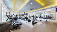 Located at 101 Zora Place in Lockhart, Florida, this fitness center offers a generously sized, single-floor layout designed to accommodate a wide range of workout routines. The space is defined by its striking architectural ceiling design — a recessed tray ceiling accented with bold yellow LED uplighting — which adds a dynamic, energetic atmosphere throughout the room. Large ceiling fans and recessed lighting provide both comfort and visibility across the entire floor.

The flooring features durable speckled rubber or epoxy-style material, suitable for heavy equipment and high-traffic use. A full-length mirror along one wall visually expands the space and supports form-checking during workouts. Natural light flows in through multiple windows, offering views of outdoor greenery and creating a bright, welcoming environment.

The cardio zone is well-stocked with treadmills, StairMaster steppers, elliptical trainers, and air bikes. The strength training area includes a power rack system, barbells, bumper plates, kettlebells, medicine balls, resistance bands, and a dedicated bench press station. A wall-mounted rack organizes free weights and accessories efficiently. A flatscreen TV is mounted near the functional training area for entertainment or instructional use. The space is thoughtfully arranged to allow fluid movement between cardio, functional, and strength training zones.