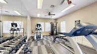 This fitness center is a single-floor facility designed to support a comprehensive workout experience. The space is bright and open, benefiting from multiple windows fitted with white horizontal blinds that allow ample natural light to fill the room. The neutral, warm-toned walls and patterned carpet tile flooring contribute to a clean and functional atmosphere.

The room is organized to accommodate both cardiovascular and strength training activities. Two True-brand treadmills are positioned toward one end of the space, accompanied by an elliptical machine and a multi-function cable and pulley resistance station. A full rack of hex dumbbells in graduated weights lines one wall, offering a wide range of options for free weight exercises. An adjustable workout bench completes the strength training setup.

A wall of floor-to-ceiling mirrors spans one side of the room, visually expanding the space and allowing users to monitor their form during exercise. Multiple ceiling fans with integrated lighting ensure comfortable air circulation throughout. Overhead fluorescent panel lighting provides consistent, even illumination.

A mounted flat-screen television is positioned for visibility across the workout area, and a fitness center rules board is displayed on the wall. A water cooler station offers added convenience. The space is accessed through elegant French doors, adding a refined architectural touch to this practical and well-appointed facility.
