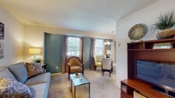 This single-floor residence offers a thoughtfully designed layout that balances comfort and functionality. The open-plan living and dining area serves as the heart of the home, characterized by plush wall-to-wall carpeting, warm neutral tones, and a striking teal accent wall that anchors the space. Multiple windows dressed with elegant draping curtains allow generous natural light to fill the room, creating a bright and welcoming atmosphere. The living area provides ample room for comfortable seating arrangements, while the adjacent dining space is illuminated by a classic chandelier, making it ideal for both everyday meals and entertaining. A wood-toned media console adds a warm, functional touch near the entryway. The kitchen is efficiently designed with light wood cabinetry, a gas range, full-size refrigerator, and tile flooring, offering practical storage and workspace. The bedroom continues the teal accent wall motif, lending a calm and restful ambiance. Two windows provide natural light and outdoor views, while carpeted flooring and warm lamp lighting contribute to a cozy retreat. The consistent use of teal accents throughout the residence creates a unified and visually appealing aesthetic that flows naturally from room to room.