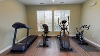 This fitness center is a thoughtfully designed, single-floor exercise facility offering a comprehensive range of workout options. The space features warm-toned, light green walls and durable wood-style flooring throughout, creating an energizing yet welcoming atmosphere. Recessed lighting complements the abundant natural light that streams in through multiple large windows fitted with blinds, offering views of the surrounding outdoor pool area and tree line beyond.

The cardio zone is equipped with a treadmill, a recumbent stationary bike, an elliptical trainer, and an upright spin bike, each placed on protective floor mats. For strength training, the facility includes a multi-function cable machine, a power tower for bodyweight exercises, and a full rack of dumbbells in varied weights. A dedicated shelf unit provides organized storage for resistance bands, hand weights, and miscellaneous accessories.

Large wall-mounted mirrors run along one side of the room, visually expanding the space and allowing users to monitor their form during workouts. Stability balls in multiple sizes are available for core and flexibility training. A wall-mounted television provides entertainment during cardio sessions. The space also features clearly posted fitness center rules and a sanitizing station near the entrance, emphasizing cleanliness and user safety throughout.