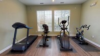 This fitness center is a thoughtfully designed, single-floor exercise facility offering a comprehensive range of workout options. The space features warm-toned, light green walls and durable wood-style flooring throughout, creating an energizing yet welcoming atmosphere. Recessed lighting complements the abundant natural light that streams in through multiple large windows fitted with blinds, offering views of the surrounding outdoor pool area and tree line beyond.

The cardio zone is equipped with a treadmill, a recumbent stationary bike, an elliptical trainer, and an upright spin bike, each placed on protective floor mats. For strength training, the facility includes a multi-function cable machine, a power tower for bodyweight exercises, and a full rack of dumbbells in varied weights. A dedicated shelf unit provides organized storage for resistance bands, hand weights, and miscellaneous accessories.

Large wall-mounted mirrors run along one side of the room, visually expanding the space and allowing users to monitor their form during workouts. Stability balls in multiple sizes are available for core and flexibility training. A wall-mounted television provides entertainment during cardio sessions. The space also features clearly posted fitness center rules and a sanitizing station near the entrance, emphasizing cleanliness and user safety throughout.