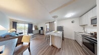 This single-floor unit offers a well-planned open-concept layout that seamlessly connects the kitchen, dining, and living areas. The kitchen is a standout feature, showcasing a granite-topped island with bar seating, light wood cabinetry, stainless steel appliances including a refrigerator, range, and microwave, all complemented by wood-look plank flooring. A striking sliding barn door serves as a stylish architectural accent, separating the main living space from the private areas of the home. The living room is bright and welcoming, with large windows dressed in dark curtain panels that allow natural light while offering privacy, and a textured ceiling that adds character overhead. The bedroom is a calm and restful retreat, featuring plush carpeting, a generously sized window with outdoor views of surrounding greenery, and ample space for furnishings. The bathroom is clean and modern, featuring a white vanity with a solid surface countertop, a large mirror, and light wood cabinetry. The overall ambiance of the space is contemporary yet warm, with a cohesive neutral palette accented by natural wood tones and modern fixtures that create an inviting and functional living environment.