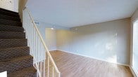 This two-story residence is designed to maximize functionality within a compact yet efficient footprint. The layout is spread across two floors, providing a natural separation between living and sleeping areas. The main living space features light-toned hardwood-style flooring that adds warmth and visual continuity throughout the room. Neutral-toned walls in soft gray and beige hues create a clean, versatile backdrop suitable for a variety of décor styles. A textured ceiling adds subtle architectural interest overhead, while recessed lighting provides practical illumination. A white-railed staircase connects the two floors, serving as a straightforward and space-conscious architectural element. Visible HVAC venting ensures year-round climate control. The overall ambiance is bright and understated, with clean lines and minimal detailing lending the space a move-in-ready appeal. The single bedroom and bathroom are thoughtfully incorporated into the layout, making this residence a well-suited option for individuals or couples seeking a manageable, low-maintenance living space across two distinct levels.