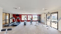 Located at 501 16th Ave N in Phenix City, Alabama, this single-floor fitness center offers a generously proportioned workout space designed to accommodate a full range of exercise needs. The room is finished with durable carpet tile flooring and a textured ceiling fitted with recessed lighting, creating a bright and functional environment. Multiple windows along the walls invite abundant natural light and provide views of the surrounding outdoor areas, enhancing the overall atmosphere of the space. A standout architectural feature is the floor-to-ceiling stone fireplace with a rustic wooden mantel, adding a distinctive and unexpected character to the fitness setting. The main workout area is divided into clearly defined zones: a cardio section anchored by treadmills, an elliptical trainer, and a stationary cycle, alongside a strength training section equipped with a multi-station cable machine, weight benches, and additional resistance equipment. A bold red accent wall with a wall-mounted television adds visual energy to the cardio zone, while the opposite side of the room maintains a neutral, lighter palette. A glass-panel door provides access to an outdoor area, further connecting the interior to the exterior environment. The layout is open and spacious, allowing for comfortable movement between equipment throughout the facility.