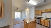 This single-floor residence is designed with an open, functional layout that connects the kitchen and living area seamlessly. The kitchen is a standout feature, equipped with stainless steel appliances including a range, microwave, refrigerator with ice maker, and dishwasher. Light wood cabinetry with ample upper and lower storage lines the walls, complemented by dark countertops and a double basin sink. A central kitchen island provides additional prep space and informal seating potential, while wood-style flooring ties the kitchen zone together.

The adjacent living area transitions to plush carpet and receives natural light through a large sliding glass door fitted with vertical blinds, opening to an outdoor space. A ceiling fan adds comfort to the room. A hallway leads to the private bedroom, which features a wide window overlooking the surrounding landscape and soft carpeted flooring.

The bathroom includes a full tub-shower combo with built-in shelving, wood-style flooring, and access to a vanity area. A generously sized walk-in closet with multi-level wire shelving and carpeted flooring provides excellent storage. Overall, the unit offers a clean, neutral palette and practical design throughout.