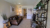 This single-floor space features a living room with a warm, inviting ambiance defined by neutral beige walls and wall-to-wall carpeting that adds a sense of comfort underfoot. The textured ceiling with crown molding detailing lends a classic, finished look to the room. A plush brown leather sofa serves as the centerpiece of the living area, complemented by decorative accent pillows, while a floor lamp provides warm ambient lighting. An ornate wrought-iron shelving unit on one side of the room offers display space for plants and decorative items, adding visual interest and a touch of greenery to the interior. Wall art, including a large leaf-motif canvas, contributes to the room's tasteful, casual aesthetic. A flat-screen television is positioned for comfortable viewing. An open doorway leads to an adjacent room, suggesting a connected and functional layout throughout the space. The overall atmosphere is cozy and well-appointed, with thoughtful décor choices that create a lived-in, homely feel suited for everyday relaxation.