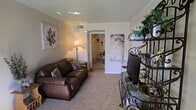 This single-floor space features a living room with a warm, inviting ambiance defined by neutral beige walls and wall-to-wall carpeting that adds a sense of comfort underfoot. The textured ceiling with crown molding detailing lends a classic, finished look to the room. A plush brown leather sofa serves as the centerpiece of the living area, complemented by decorative accent pillows, while a floor lamp provides warm ambient lighting. An ornate wrought-iron shelving unit on one side of the room offers display space for plants and decorative items, adding visual interest and a touch of greenery to the interior. Wall art, including a large leaf-motif canvas, contributes to the room's tasteful, casual aesthetic. A flat-screen television is positioned for comfortable viewing. An open doorway leads to an adjacent room, suggesting a connected and functional layout throughout the space. The overall atmosphere is cozy and well-appointed, with thoughtful décor choices that create a lived-in, homely feel suited for everyday relaxation.
