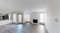 This single-floor residence offers a clean, modern aesthetic throughout, with light neutral walls and durable wood-look flooring that flows seamlessly across the main living areas. The spacious living room is a standout feature, anchored by a crisp white fireplace and bathed in natural light from multiple windows fitted with horizontal blinds. A glass-paneled exterior door provides convenient outdoor access directly from the living space, enhancing the open, airy feel of the room.

The kitchen is practical and well-designed, featuring gray stone-look countertops, a stainless steel sink, and a stainless steel refrigerator. A pass-through counter connects the kitchen to the adjacent dining or living area, promoting an open flow throughout the home. Generous windows in the kitchen area flood the space with additional natural light.

The bedroom is a comfortable, well-proportioned room with the same wood-look flooring, a ceiling fan with lighting, and a window for natural light and ventilation.

The bathroom is thoughtfully appointed with gray shaker-style cabinetry, a gray countertop vanity, dual mirrors, brushed nickel fixtures, and ample overhead cabinet storage. A separate tub and shower combination with a curved curtain rod and brushed nickel hardware completes the bathroom. Clean white walls and warm wood-look flooring tie the space together with a cohesive, contemporary style.