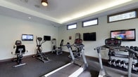 This fitness center offers a practical and well-appointed space designed to support a variety of workout routines. The room features a modern aesthetic with smooth, light-grey walls and a subtle cove lighting system along the ceiling perimeter that provides warm, ambient illumination throughout the space. A ceiling fan adds to the comfort of the environment during exercise sessions.

The facility is equipped with a selection of cardio machines, including treadmills and an elliptical trainer, as well as dedicated strength training equipment such as a cable machine and a seated resistance unit. The equipment is arranged to allow comfortable movement and access throughout the room. Durable, dark rubber flooring covers the entire area, providing both safety and noise reduction during workouts.

Multiple wall-mounted flat-screen televisions are positioned at convenient viewing angles, allowing users to stay entertained or informed during exercise. A row of horizontal windows near the ceiling allows natural light to enter the space while maintaining privacy. The overall ambiance is clean, focused, and functional, making it a well-suited environment for daily fitness activity.