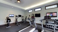 This fitness center offers a practical and well-appointed space designed to support a variety of workout routines. The room features a modern aesthetic with smooth, light-grey walls and a subtle cove lighting system along the ceiling perimeter that provides warm, ambient illumination throughout the space. A ceiling fan adds to the comfort of the environment during exercise sessions.

The facility is equipped with a selection of cardio machines, including treadmills and an elliptical trainer, as well as dedicated strength training equipment such as a cable machine and a seated resistance unit. The equipment is arranged to allow comfortable movement and access throughout the room. Durable, dark rubber flooring covers the entire area, providing both safety and noise reduction during workouts.

Multiple wall-mounted flat-screen televisions are positioned at convenient viewing angles, allowing users to stay entertained or informed during exercise. A row of horizontal windows near the ceiling allows natural light to enter the space while maintaining privacy. The overall ambiance is clean, focused, and functional, making it a well-suited environment for daily fitness activity.