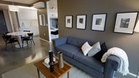 This single-floor space showcases a thoughtfully designed open-concept layout that blends the living area, kitchen, and workspace into one cohesive environment. The living room is anchored by a contemporary charcoal-blue tufted sofa, complemented by a warm wood coffee table and a patterned area rug atop polished concrete floors — a combination that balances industrial and modern aesthetics. Rich, earthy taupe walls lend warmth and depth to the space, while a trio of framed black-and-white artwork adds a curated, gallery-like quality. A built-in white shelving unit serves as a functional and visual transition between the living area and an intimate workspace, which features a sleek built-in desk and stylish seating. The adjacent kitchen is finished with clean white cabinetry and a dark tile backsplash, contributing to the space's contrast-driven design palette. A wall-mounted HVAC unit ensures climate comfort, while the polished concrete flooring throughout ties the entire space together with a cohesive, contemporary feel. The overall ambiance is modern and sophisticated, with a neutral color palette that promotes both focus and relaxation.