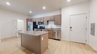 This contemporary single-floor unit offers a clean, modern aesthetic throughout, defined by light wood-style flooring, crisp white walls, and an open, functional layout. The kitchen is a standout feature, equipped with a full suite of stainless steel appliances — including a gas range, over-the-range microwave, French door refrigerator with ice maker, and dishwasher. Warm-toned wood cabinetry extends to the ceiling for generous storage, while granite countertops and a central island provide ample prep and dining space. The living area is bright and open, with a ceiling fan and access to a large street-facing entry with floor-to-ceiling windows that invite abundant natural light and views of the surrounding streetscape. The bedroom is generously sized, carpeted for comfort, and features multiple large windows with blinds, allowing for excellent light control. A walk-in closet connects directly to a full bathroom fitted with a soaking tub and shower combination, a granite-topped wood vanity, and light wood flooring. A separate half bathroom with a matching vanity and grab bar offers additional convenience. Throughout, the unit maintains a cohesive, contemporary design with quality finishes and a smart, well-organized floor plan.