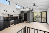 Located along scenic Laguna Canyon Road in Laguna Beach, CA, this contemporary two-level loft showcases a striking industrial-modern aesthetic with thoughtfully curated details throughout. The main level greets visitors with soaring ceilings accented by exposed structural beams painted white, polished concrete floors, and a dramatic wall of black-framed floor-to-ceiling windows that flood the space with natural light and frame lush canyon greenery. Track lighting and a ceiling fan complete the airy, open atmosphere.

The kitchen is outfitted with dark matte cabinetry, a bold veined stone backsplash, stainless and black appliances, and a floating wood shelf with wall-mounted sconce lighting. Clerestory windows above the kitchen draw in additional natural light. A sliding door leads to a stylishly tiled bathroom featuring floor-to-ceiling dark brick-style mosaic tile walls, a walk-in shower, a wall-mounted toilet, and hexagonal penny tile flooring.

An industrial steel ladder and a separate open staircase with black metal railings lead to the mezzanine loft level above, which offers an intimate overlook of the main space below. French doors with frosted glass panels open to an additional flexible area on the upper level. The overall design balances raw industrial elements with refined finishes, creating a versatile and visually compelling space.