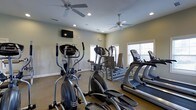 This single-story fitness center offers a practical and comfortable environment for cardio-focused workouts. The space is outfitted with multiple elliptical trainers, treadmills, and a cable weight machine, providing a solid range of equipment for residents or members. Neutral-toned walls and carpeted flooring give the room a clean, understated aesthetic throughout.

Natural light filters in through several windows fitted with horizontal blinds, brightening the space and creating an open, airy atmosphere. Two ceiling fans are evenly distributed across the room, helping to maintain air circulation during workouts. Recessed lighting supplements the natural light, ensuring the space remains well-lit regardless of the time of day.

A wall-mounted flat-screen television is positioned at the front of the room, offering entertainment during exercise sessions. The layout is thoughtfully arranged to allow comfortable movement between machines without overcrowding. Overall, the fitness center delivers a functional and inviting setting well-suited for daily cardio training.