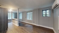 This single-floor unit offers a thoughtfully designed open-concept layout that seamlessly connects the living, dining, and kitchen areas. The expansive main living space is finished with rich hardwood-style flooring and neutral gray walls, creating a clean and contemporary aesthetic throughout. Multiple windows fitted with vertical blinds allow natural light to filter in while providing views of surrounding greenery, enhancing the overall sense of openness.

The kitchen is a standout feature, boasting sleek two-tone cabinetry in light wood and dark finishes, stainless steel appliances including a range and microwave, a tile backsplash, and a generous marble-style countertop peninsula that doubles as a breakfast bar. The open pass-through design connects the kitchen directly to the living area, making the space ideal for both daily living and entertaining.

The bedroom is a well-proportioned private retreat, complete with hardwood-style flooring, a large window overlooking lush greenery, and a wall-mounted split air conditioning unit for personalized climate control. The bathroom is neatly appointed with tile flooring, white wall tile wainscoting, a glass-enclosed shower, vanity with overhead cabinetry, and clean, modern fixtures. Together, these spaces form a cohesive and functional unit with a fresh, contemporary finish.