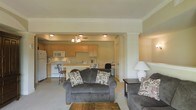 This single-floor space is designed around an open-concept layout that seamlessly connects the living area and kitchen, creating a cohesive and functional environment. The living area is furnished with plush grey upholstered seating, including a loveseat and armchair accented with patterned throw pillows, arranged around a rustic wooden coffee table. A large table lamp provides warm ambient lighting, complementing the neutral beige and cream tones of the walls and carpeted flooring. Crown molding adds a subtle architectural detail throughout. The kitchen, visible from the living area, features light wood cabinetry, white appliances including a refrigerator, microwave, and range, and a breakfast bar with barstool seating — ideal for casual dining. A small chandelier overhead adds a touch of warmth to the kitchen space. A structural column subtly defines the transition between the two areas while maintaining the open feel. The overall ambiance is warm, practical, and inviting, with a consistent neutral palette and functional furnishings that make efficient use of the available space.