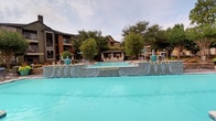 This single-story community space offers a well-rounded outdoor environment designed for recreation and gathering. The centerpiece is a generously sized resort-style swimming pool featuring striking blue mosaic tile accents along its raised water feature wall, creating a vibrant and inviting atmosphere. The pool area is complemented by a covered gazebo or cabana structure, providing shaded seating and a comfortable retreat for guests. Surrounding the pool, mature trees offer natural shade and a lush backdrop, with multi-story residential buildings visible in the distance.

The main building is clad in vertical panel siding in a neutral cream tone with brown trim accents, topped by a dark shingled roof. Multiple windows line the exterior, allowing natural light into the interior while maintaining visibility to the outdoor surroundings. The building features at least two exterior access doors and is flanked by a spacious concrete patio area.

Adjacent to the building, the outdoor grounds include picnic tables sheltered by colorful umbrellas, a playground structure, and open paved areas suitable for casual outdoor activities. The combination of the pool, shaded seating, recreational equipment, and functional building makes this a versatile community facility well-suited for a variety of uses and gatherings.