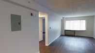 This single-floor unit offers a clean, functional layout designed for comfortable everyday living. The space is anchored by rich, dark hardwood flooring that runs throughout, providing warmth and continuity from room to room. Neutral light gray walls create a calm, versatile backdrop that adapts well to a variety of furnishing styles.

The main living area benefits from a large window that invites natural light into the space, with a wall-mounted HVAC unit positioned beneath it for year-round climate control. A structural beam accent along the ceiling adds architectural character to the open layout.

A doorway visible from the main living area leads into a secondary room, also finished with hardwood flooring and similarly neutral wall tones, maintaining a cohesive aesthetic throughout. The transition between spaces is defined by a clean white door frame, reinforcing the crisp, well-maintained character of the interior.

Safety features including smoke and alarm detectors are installed on the ceiling and walls. The overall ambiance is bright, tidy, and move-in ready, reflecting a straightforward and practical design sensibility suited for a range of occupants.