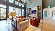 Located at 2370 Sever Rd in Lawrenceville, GA, this beautifully designed community clubhouse offers a sophisticated and welcoming atmosphere across its single-floor layout. The entry foyer immediately impresses with soaring double-height ceilings, bold hexagonal tile flooring, and an elegant glass-railed staircase that adds a modern architectural accent.

The main lounge area is a standout, featuring dramatic floor-to-ceiling windows that flood the space with natural light and offer expansive outdoor views. A coffered ceiling with an open grid detail adds architectural depth, while plush seating arrangements — including a large sectional sofa and richly upholstered armchairs — surround a built-in entertainment wall with marble-accented shelving.

Adjacent to the lounge is a well-equipped bar and social area, highlighted by a long marble-topped island with green velvet bar seating, pendant lighting, and backlit open shelving. A wall-mounted TV, wet bar sink, and built-in wine refrigerator make this an ideal gathering space. Wood-look tile flooring throughout this zone complements the warm, contemporary aesthetic.

Additional functional areas include a corridor with a commercial-grade glass-door refrigerator, elevator access, and curated decorative shelving. The overall design balances modern elegance with inviting comfort, making it a premier amenity space.