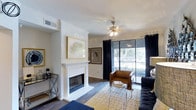 Located in Arlington, this single-story space presents a thoughtfully designed living area that balances comfort and modern style. The living room serves as the centerpiece, highlighted by a classic white-mantled fireplace that anchors the space with a sense of warmth and elegance. Crown molding frames the white ceiling, adding a refined architectural detail, while a ceiling fan with a contemporary chrome fixture ensures year-round comfort.

The flooring throughout features rich hardwood-style planks that contrast beautifully with the crisp white walls. Framed artwork adorns the walls, contributing to the cultivated aesthetic of the space. A large sliding glass door dressed with floor-to-ceiling dark curtains floods the room with natural light and opens to a balcony, offering views of surrounding mature trees and greenery.

The open layout accommodates a living arrangement with ample room for seating and a coffee table area. A bar cart station is neatly tucked near the wall, adding a functional yet stylish touch. Overall, the space exudes a polished, inviting ambiance suited for comfortable everyday living.