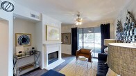 Located in Arlington, this single-story space presents a thoughtfully designed living area that balances comfort and modern style. The living room serves as the centerpiece, highlighted by a classic white-mantled fireplace that anchors the space with a sense of warmth and elegance. Crown molding frames the white ceiling, adding a refined architectural detail, while a ceiling fan with a contemporary chrome fixture ensures year-round comfort.

The flooring throughout features rich hardwood-style planks that contrast beautifully with the crisp white walls. Framed artwork adorns the walls, contributing to the cultivated aesthetic of the space. A large sliding glass door dressed with floor-to-ceiling dark curtains floods the room with natural light and opens to a balcony, offering views of surrounding mature trees and greenery.

The open layout accommodates a living arrangement with ample room for seating and a coffee table area. A bar cart station is neatly tucked near the wall, adding a functional yet stylish touch. Overall, the space exudes a polished, inviting ambiance suited for comfortable everyday living.