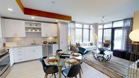 This contemporary single-floor residence showcases a thoughtfully designed open-concept layout that seamlessly integrates the kitchen, dining, and living areas into one cohesive and inviting space. The kitchen is outfitted with crisp white cabinetry, granite countertops, stainless steel appliances, and an open shelving display — all anchored by a bold burnt-orange accent wall that adds a vibrant splash of color. Adjacent to the kitchen, the dining area features a round glass-top table paired with sleek modern chairs, creating an intimate setting ideal for entertaining. The living room is the centerpiece of the unit, bathed in abundant natural light streaming through expansive floor-to-ceiling windows that frame striking urban city views. The space is furnished with a contemporary sofa, eclectic accent chairs, and layered rugs that add warmth and texture to the light hardwood flooring. Recessed lighting throughout complements the natural light, while the open layout enhances the sense of spaciousness. The overall ambiance strikes a balance between modern sophistication and comfortable livability, making it an ideal urban retreat.