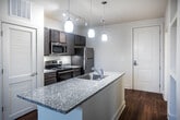 This thoughtfully designed one-bedroom, one-bathroom residence in Memphis, Tennessee offers a functional and stylish single-floor layout. The kitchen serves as a standout feature, boasting rich dark-stained cabinetry, granite countertops, and a full suite of stainless steel appliances including an electric range, built-in microwave, and refrigerator. A peninsula with a granite countertop and integrated sink provides additional prep space and casual seating, while pendant lighting adds a contemporary touch overhead. Track lighting illuminates the kitchen area efficiently. Warm hardwood-style flooring runs through the kitchen and main living areas, creating a cohesive and inviting atmosphere. The bedroom is carpeted for comfort and receives natural light through a pair of windows fitted with blinds. A spacious walk-in closet with wire shelving and additional hanging storage offers generous organizational capacity. The bathroom features matching dark cabinetry with a granite-topped vanity, a large mirror, hardwood-style flooring, and a shower-tub combination. The overall layout, as seen in the dollhouse view, is compact yet efficiently arranged, making excellent use of every square foot in this Memphis residence.