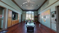 This single-floor community center is thoughtfully designed to serve a variety of programs and services. The centerpiece of the space is a generously sized main hall characterized by soaring ceilings accented with crown molding and warm teal-painted walls. Rich, dark hardwood floors extend throughout, lending a polished and welcoming ambiance. A large arched window at the far end of the main hall floods the room with abundant natural light and offers views of the outdoor surroundings, while recessed lighting and a ceiling fan ensure year-round comfort. A central activity table with seating anchors the room, and a bookshelf stocked with resources lines one wall alongside a dedicated "Social Services" display board. Elegant French doors with glass pane detailing connect the main hall to adjacent areas, creating an open, interconnected flow. A kitchenette is accessible from the main area, outfitted with warm wood cabinetry, pendant lighting, and a full complement of appliances, providing practical support for events and programs. A separate children's program room features its own tall arched window, round activity tables, and dedicated workstations, making it a bright and functional space for youth-focused activities. Together, the rooms create a versatile, community-oriented environment designed for engagement and accessibility.
