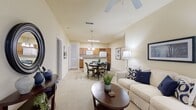This single-story unit offers a thoughtfully arranged floor plan that maximizes its compact footprint. The open-concept main living area seamlessly connects the living room, dining area, and kitchen, creating a cohesive and functional space. The living room features vaulted ceilings with a ceiling fan, soft neutral walls, plush carpeting, and a private entrance door with a sidelight window that brings in natural light. The kitchen is equipped with warm wood cabinetry, a built-in wine rack, granite-style countertops, a breakfast bar peninsula, white appliances including a refrigerator and microwave, and a classic checkered tile floor. Adjacent to the kitchen, the dining area accommodates a four-person table beneath a decorative pendant chandelier. A hallway leads to the private bedroom, which features vaulted ceilings, a ceiling fan, carpet flooring, and multiple windows with drape treatments. A dedicated closet provides additional storage. The full bathroom includes a vanity with granite-style countertops, a tub-shower combination, and matching checkered tile flooring consistent with the kitchen. Overall, the unit presents a warm and cohesive aesthetic throughout.