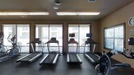 This fitness center is a purpose-built exercise facility designed to support a well-rounded workout routine. The space is anchored by a row of treadmills, each equipped with individual mounted screens, allowing users to stay entertained or track their progress during cardio sessions. Additional cardio equipment, including a stationary bike, is also present, offering variety for different training preferences.

Two large windows span the far wall, fitted with horizontal blinds and providing a generous view of the surrounding exterior — an outdoor area with additional building structures visible beyond. The natural light filtering through these windows contributes to an open, energizing atmosphere within the space.

Overhead, fluorescent lighting ensures the room is evenly and brightly lit regardless of the time of day. A ceiling fan is mounted centrally, promoting air circulation throughout the workout area. The flooring is composed of durable, dark-toned material well-suited for high-traffic fitness use.

The walls are finished in a warm neutral tone, and the overall layout is clean and organized, making navigation through the space straightforward. The single-floor design ensures full accessibility throughout. This fitness center offers a practical and comfortable environment for daily exercise.