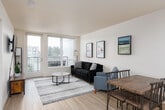 This contemporary single-floor residence is designed with a clean, modern aesthetic throughout. The open-concept layout connects the kitchen, dining, and living areas seamlessly, creating a spacious and functional flow. Light wood-style flooring runs through the main living spaces, complementing the crisp white walls and recessed lighting overhead.

The kitchen features a generous peninsula with a white countertop and built-in sink, providing ample prep space and casual bar seating. Dark espresso cabinetry offers substantial storage, while stainless steel appliances — including a refrigerator, range, and over-the-range microwave — complete the functional setup.

The living area is bright and welcoming, anchored by large sliding glass doors that lead to a private balcony. These expansive windows flood the room with natural light and offer views of the surrounding neighborhood.

The bedroom is a serene retreat, carpeted for comfort, and features a large window with outdoor views, as well as direct access to a well-organized walk-in closet with built-in wire shelving.

The bathroom is finished with light wood-look flooring, a dark-toned vanity with a white countertop sink, and a clean, efficient layout. Overall, the unit presents a cohesive, contemporary design with practical features throughout.