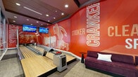 Located in West Palm Beach, FL, this dynamic single-floor entertainment and game room delivers a bold, energetic atmosphere across a generously sized footprint. The space is anchored by a striking bowling alley wrapped in vivid red graphic murals featuring bold bowling-themed typography such as "Cranker," "Stroker," and "Strike," accompanied by overhead monitors for score tracking. Curved purple velvet seating runs alongside the lanes, providing a stylish spectator area. Adjacent to the bowling section, a dedicated karaoke pod features a curved built-in bench, ambient purple LED cove lighting, a wall-mounted TV, and a tufted ottoman centerpiece — creating an intimate, lounge-like enclave. The main open game area includes a full-size billiards table and a ping-pong table beneath large industrial-style pendant lights. Floor-to-ceiling windows along one wall flood this zone with natural light and offer views of a lush outdoor pool area lined with palm trees. Additional seating nooks with orange accent chairs and low-profile sofas provide flexible social spaces throughout. Recessed lighting and a dark tray ceiling unify the design, while glass partitions between zones maintain an open, connected feel throughout this entertainment-rich environment.