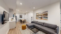 This single-floor space showcases a cohesive modern design throughout, anchored by light hardwood flooring and crisp white walls that create a bright, airy atmosphere. The open-plan living and dining area serves as the heart of the unit, featuring recessed lighting, a large sectional sofa, and a minimalist coffee table set atop a woven area rug. Bold, colorful abstract artwork adds visual warmth to the living space. Glass block windows along one wall allow diffused natural light to filter through, complementing the wall-mounted television near the entry.

The kitchen is efficiently designed with floor-to-ceiling taupe cabinetry, white quartz countertops, a subway tile backsplash, under-cabinet lighting, and stainless steel appliances including a French door refrigerator and dishwasher. The dining area accommodates a natural wood table with seating, positioned near louvered closet doors for additional storage.

The primary bathroom is a standout feature, presenting a freestanding soaking tub, a floating wood-toned vanity with a black matte faucet, large-format tile flooring, and a dramatic circular pendant light. A second bathroom mirrors the modern finishes with a floating vanity and shower. The bedrooms are serene and minimalist, with large windows framing outdoor views.