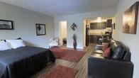 This single-floor studio unit offers an open-concept layout that efficiently combines the sleeping, living, and kitchen areas into one cohesive space. The flooring throughout is a warm hardwood-style material that adds a sense of continuity across the entire unit.

The main living area accommodates both a bed and a seating arrangement, with rich burgundy area rugs anchoring each zone and adding color contrast against the neutral gray walls. A textured ceiling runs throughout the space, and a decorative tall vase with dried branches serves as a subtle design accent near the hallway entrance.

The kitchen is positioned toward the rear of the unit and features dark espresso-toned cabinetry paired with a black refrigerator and a gas range. A small dining area with a table and chairs is tucked neatly alongside the kitchen, maximizing the use of available space.

A hallway visible from the main room leads to the bathroom, which appears to offer standard fixtures. The overall ambiance is cozy and functional, with a straightforward layout designed to make practical use of every square foot available in this compact single-room living space.