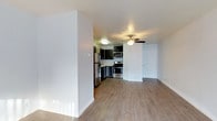 This single-floor residence offers a clean, open-concept layout that seamlessly connects the living area and kitchen, creating a functional and inviting atmosphere. The living space features warm hardwood-style flooring that extends throughout, bright white walls, and a ceiling fan that enhances air circulation and comfort. A partial dividing wall adds a subtle sense of definition between the living and kitchen areas while maintaining the open feel of the space. The kitchen is well-appointed with dark cabinetry that provides a striking contrast against the lighter surroundings. Stainless steel appliances — including a refrigerator, stove, oven, and microwave — lend a modern, polished look to the cooking space. The cabinetry offers ample storage, and the layout is designed for practical everyday use. The living area benefits from generous open floor space, making it highly versatile for various furniture arrangements. Crisp white baseboards and neutral-toned walls contribute to the clean, contemporary aesthetic throughout. A white paneled door is visible at the far end of the space, hinting at additional rooms or storage beyond. Overall, this space presents a tidy, move-in-ready interior with a cohesive modern design.