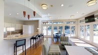 This single-floor community amenity space is thoughtfully designed to offer a welcoming and versatile gathering environment. The open-concept layout seamlessly connects a bar area, casual lounge, and dining zone, creating a fluid and functional space for residents and guests alike.

The bar area features a granite countertop with sleek bar seating, accompanied by stylish pendant lighting with warm amber tones that add a distinctive decorative touch. Flush-mount drum fixtures supplement the recessed lighting throughout, ensuring the space is well-lit and inviting at all times.

Rich hardwood flooring runs throughout the main area, lending warmth and continuity to the design. The lounge section is furnished with striped upholstered seating, offering a comfortable and casual retreat, while a separate dining area provides a more structured setting with round tables and dark wood chairs.

Floor-to-ceiling glass doors and a generous bank of transom windows line the rear wall, inviting abundant natural light and offering unobstructed views of the outdoor pool and surrounding greenery. The neutral color palette of soft whites and warm grays throughout the interior complements the natural tones visible beyond, creating a seamless indoor-outdoor connection. Overall, the space strikes a balance between refined aesthetics and practical, everyday usability.
