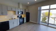 This single-floor space showcases a thoughtfully designed open-concept kitchen and living area. The kitchen features grey shaker-style cabinetry with sleek hardware, complemented by light granite countertops and a white subway tile backsplash that adds a clean, classic touch. Black appliances, including an electric range, built-in microwave, and refrigerator, provide a striking contrast against the lighter cabinetry. A stainless steel sink is positioned beneath upper cabinets, maximizing functionality. A wall-mounted range hood adds a practical yet stylish focal point above the range. The space transitions seamlessly into an open living area, finished with light-toned laminate wood flooring that runs throughout, lending warmth and continuity to the layout. Neutral grey walls create a versatile backdrop throughout. A large sliding glass door floods the space with natural light and provides direct access to a private balcony, offering views of the surrounding greenery. Recessed lighting in the kitchen ensures ample illumination, while the overall design maintains a clean, modern aesthetic suited for everyday living.