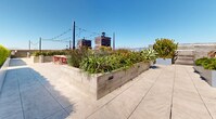 This expansive rooftop terrace offers a well-appointed outdoor retreat above the city skyline. The space is thoughtfully divided into distinct functional zones, each designed for comfort and entertaining. Large raised concrete planters filled with drought-tolerant agaves, ornamental grasses, and colorful flowering plants serve as natural dividers throughout the terrace, adding vibrant greenery against the urban backdrop.

A dedicated dining area features a rustic wood communal table with red metal chairs, positioned alongside a built-in outdoor kitchen equipped with a stainless steel grill — ideal for al fresco gatherings. String lights crisscross overhead, lending the space a warm ambiance after dark.

A separate lounge zone provides a more intimate setting, centered around a round fire pit surrounded by plush, cushioned wicker sofas and accent chairs. A wall-mounted screen adds a versatile entertainment element to this seating area.

Manicured hedgerows line portions of the perimeter, offering both privacy and a refined aesthetic, while open sightlines deliver panoramic views of the surrounding city skyline and distant hills. The entire terrace is finished with large-format porcelain tile pavers, lending a clean, contemporary feel to the overall design.