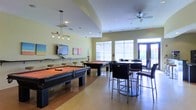 This single-floor recreational lounge is designed to serve as a social and entertainment hub. The space is divided into distinct activity zones that coexist within an open, flowing layout. Two full-size pool tables with rich orange felt surfaces dominate the central area, positioned to allow ample room for play on all sides. A wall-mounted flat-screen television is installed along one wall, accompanied by a row of bar-height stools, creating a comfortable viewing area. Track lighting and a modern pendant fixture illuminate the pool tables, while a ceiling fan overhead aids in air circulation throughout the room. The walls are painted in a soft yellow-green tone, contributing to a relaxed and inviting atmosphere. On the opposite side of the room, high-top tables paired with contemporary bar chairs provide additional seating for casual gathering. Large windows fitted with horizontal blinds line the walls, allowing generous natural light to filter into the space. A set of glass-panel doors at the far end opens to an outdoor area, enhancing the sense of openness. The polished concrete flooring ties the entire space together with a clean, modern finish.