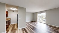 This single-floor unit offers a thoughtfully designed layout that balances open living space with functional room separation. The main living area is finished with light, wood-style laminate flooring that extends throughout, complemented by neutral gray walls and a textured ceiling. A large sliding glass door floods the room with natural light and opens onto a private balcony, offering views of the surrounding outdoor environment. The open flow between the living area and kitchen makes the space feel expansive and connected. The kitchen is equipped with rich, dark-stained wood cabinetry providing ample storage, granite-style countertops, a double stainless steel sink with a modern faucet, and black appliances including a dishwasher and refrigerator. The bedroom features plush wall-to-wall carpeting, a window with blinds for natural light and privacy, and clean neutral tones that create a calm, restful atmosphere. The bathroom includes a full tub and shower combination, a toilet, and a vanity with granite-style countertop, all finished with wood-style flooring that ties the space together. Throughout the unit, clean white trim, consistent neutral tones, and practical finishes contribute to a cohesive and comfortable living environment.