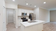 This well-appointed single-floor residence offers a clean, modern aesthetic characterized by a neutral color palette, crown molding, and light wood-style flooring throughout. The open-concept kitchen is a standout feature, centered around a large island with a built-in sink and bright white quartz countertops. White shaker-style cabinetry lines the walls, complemented by a subway tile backsplash and a full suite of stainless steel appliances, including a refrigerator, electric range, and over-the-range microwave. The kitchen flows seamlessly into a generous living and dining area, which is bathed in natural light from multiple windows dressed with horizontal blinds. A stylish chandelier and a ceiling fan with integrated lighting add character and comfort to the open living space. The bathroom features light wood-style flooring, a white vanity with ample storage, a glass-enclosed shower, and a separate soaking tub with a molded surround. Recessed lighting throughout the home ensures bright, even illumination in every room. The residence also includes an attached garage with an automatic door opener, providing convenient and secure parking or additional storage space. The overall ambiance is fresh, clean, and move-in ready.