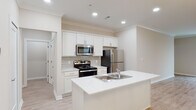 This well-appointed single-floor residence offers a clean, modern aesthetic characterized by a neutral color palette, crown molding, and light wood-style flooring throughout. The open-concept kitchen is a standout feature, centered around a large island with a built-in sink and bright white quartz countertops. White shaker-style cabinetry lines the walls, complemented by a subway tile backsplash and a full suite of stainless steel appliances, including a refrigerator, electric range, and over-the-range microwave. The kitchen flows seamlessly into a generous living and dining area, which is bathed in natural light from multiple windows dressed with horizontal blinds. A stylish chandelier and a ceiling fan with integrated lighting add character and comfort to the open living space. The bathroom features light wood-style flooring, a white vanity with ample storage, a glass-enclosed shower, and a separate soaking tub with a molded surround. Recessed lighting throughout the home ensures bright, even illumination in every room. The residence also includes an attached garage with an automatic door opener, providing convenient and secure parking or additional storage space. The overall ambiance is fresh, clean, and move-in ready.
