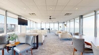 This elevated single-floor space offers a remarkable blend of modern design and breathtaking waterfront panoramas. Floor-to-ceiling windows wrap around the expansive lounge area, flooding the interior with natural light and framing sweeping views of the water and city skyline beyond. The drop-tile ceiling with recessed lighting and ceiling fans complements the open, airy layout while maintaining a clean, polished aesthetic throughout.

The main lounge is thoughtfully organized into distinct seating zones, each with its own character. Plush cream sofas anchor a media area complete with a wall-mounted flat-screen television, while clusters of contemporary armchairs and occasional tables create intimate conversation nooks. A central dining and gathering area features sleek bistro-style tables and upholstered chairs, ideal for meetings or social events. A patterned blue-grey carpet ties the space together, lending a coastal sensibility that echoes the waterfront setting.

The fully equipped kitchen features white cabinetry with frosted glass accents, a subway tile backsplash, stainless steel appliances including a full-size refrigerator, oven, and built-in microwave, and generous counter space. Large windows in the kitchen also capture the waterfront view, making it a bright and functional workspace for catering or everyday use. Overall, this space is well-suited for private events, corporate gatherings, or upscale social functions.