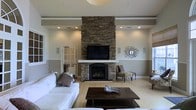 This single-floor residence offers a thoughtfully designed interior centered around a dramatic stacked-stone fireplace that serves as the focal point of the main living area. The fireplace rises from floor to ceiling, framing a mounted flat-screen television and a classic hearth below, blending rustic character with modern functionality. The living room is finished with light hardwood flooring that extends throughout, contributing to a warm and cohesive aesthetic. Recessed lighting is strategically placed across the ceiling, providing even, ambient illumination that complements the natural tones of the space. Crown molding and a wainscoting-style lower wall treatment add architectural refinement, while built-in speakers suggest the room is designed with entertainment in mind. The neutral color palette — soft whites, warm grays, and natural wood tones — lends the space a calm and inviting ambiance. A substantial dark wood coffee table anchors the seating area, and accent chairs positioned near the fireplace offer additional comfort. Decorative wall art and carefully placed accents throughout reflect a curated, tasteful approach to interior design. Overall, this home presents a harmonious balance of comfort, style, and architectural detail.