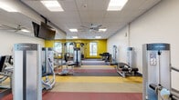 This single-floor fitness center is thoughtfully designed to support a comprehensive workout experience. The space is defined by its striking multi-colored chevron and striped carpet tile flooring in shades of yellow, red, gray, and beige, adding a dynamic and energetic visual character throughout. Bold yellow accent walls punctuate the interior, contributing to the facility's vibrant, motivating ambiance.

The cardio zone features multiple treadmills and elliptical machines positioned along floor-to-ceiling windows, offering views of the surrounding outdoor environment and allowing generous natural light to fill the space. Ceiling fans installed throughout ensure comfortable air circulation during workouts.

The strength training area is extensively equipped, housing a full rack of dumbbells and kettlebells, multiple cable and selectorized resistance machines by Life Fitness, a power rack with barbell and weight plates, and adjustable benches. Wall-mounted mirrors extend across key sections of the gym, enhancing the sense of openness and allowing users to monitor their form.

Wall-mounted flat-screen televisions are positioned for visibility across multiple workout zones. A dedicated entry area features a dual water fountain station and glass-walled corridor access, providing a clean and functional transition into the facility. Recessed panel lighting and a drop ceiling complete the polished, professional interior finish.